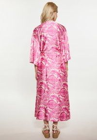 Un lungo kimono rosa con un motivo floreale paisley, realizzato in tessuto morbido; presenta maniche a kimono e una cintura da legare in vita, abbinato a sandali con zeppa color beige.