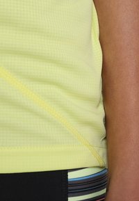 Top deportivo amarillo claro con una tela texturizada, que presenta costuras diagonales y un diseño ajustado. Cintura negra y rayada visible por debajo.