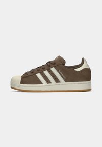 Sneaker in suede marrone con punta in gomma, tre strisce bianche e suola color crema. Presenta lacci e dettagli marroni sul collo.