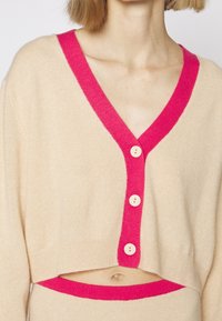 Beige lyhyt neuletakki, jossa on syvä V-pääntie, kirkkaan vaaleanpunainen koriste ja neljä kermaista nappia. Pehmeä rakenne, suorat reunat ja minimaalinen muotoilu.