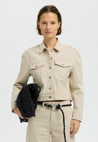 Chaqueta de mezclilla beige con un diseño recortado, cierre de botones en la parte delantera, dos bolsillos en el pecho y una textura sutil, combinada con un bolso de malla negra.