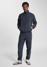 Marineblauw trainingspak met witte piping; het jack met volledige ritssluiting heeft elastische manchetten en zijzakken, en is gecombineerd met bijpassende tapered broek. Casual sportieve stijl.