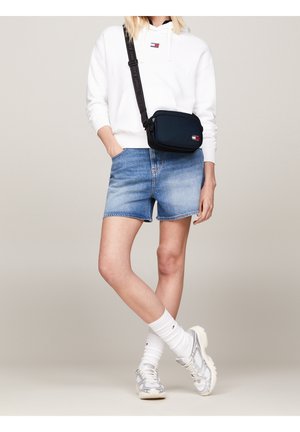 Sweat à capuche blanc avec un logo, short en jean et baskets grises. Le sac bandoulière est bleu marine avec un écusson logo et une sangle contrastante.