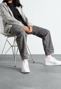 Grå zip-up huvtröja över en svart tröja, i kombination med grå jeans och vita sneakers, sittande på en metallstol mot en ljus bakgrund.