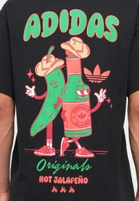 T-shirt noir avec un graphique d'un poivron vert et d'une bouteille avec des yeux, tous deux portant des chapeaux, avec le texte "ADIDAS" et "HOT JALAPEÑO" en couleurs vives.