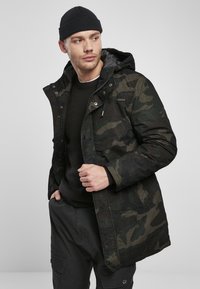 Parka z vojaškim vzorcem v temno zeleni in črni barvi, ki ima kapuco z vrvico, zadrgo in teksturiran material. Nosi se preko črnepuloverja.