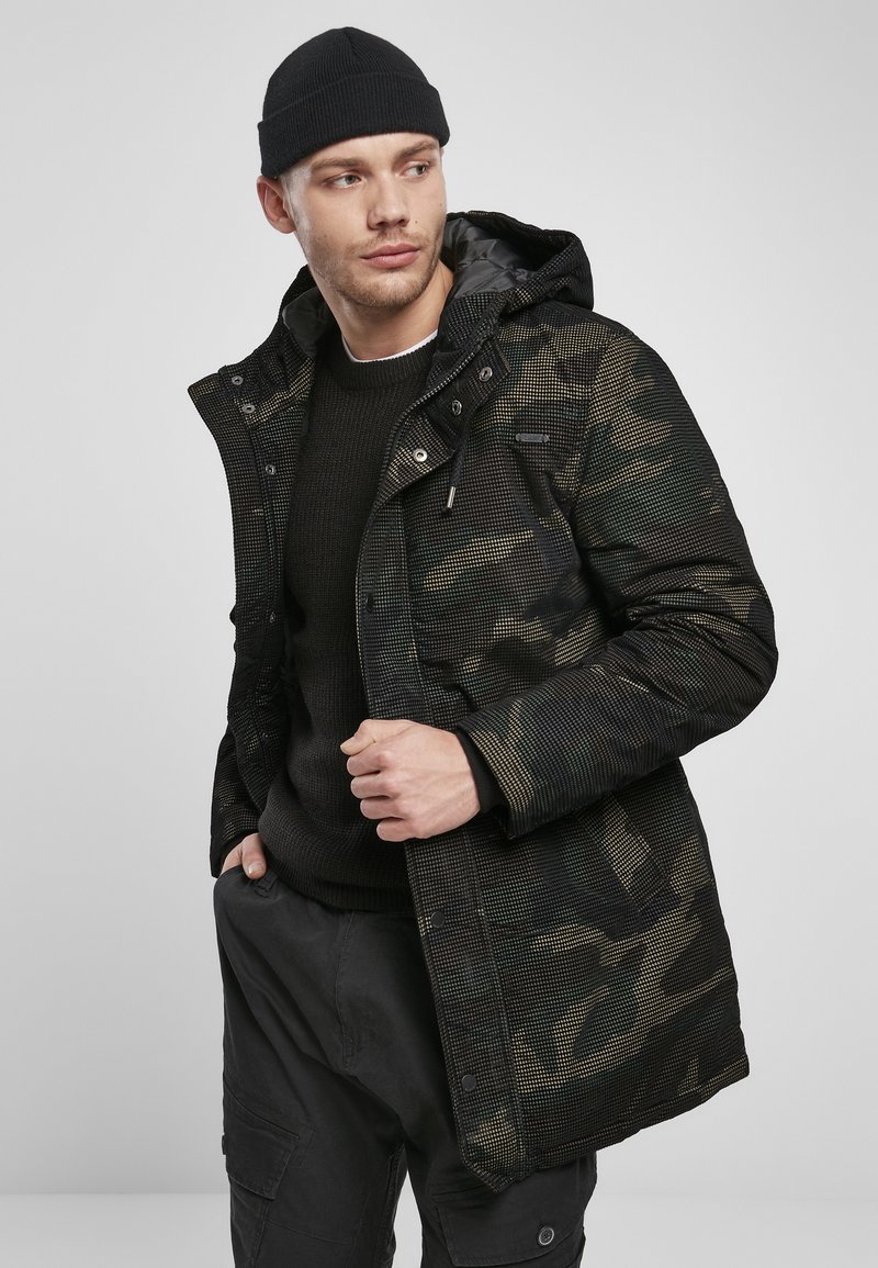 Parka z vojaškim vzorcem v temno zeleni in črni barvi, ki ima kapuco z vrvico, zadrgo in teksturiran material. Nosi se preko črnepuloverja.