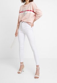 Vrouw draagt een roze sweatshirt met rood-witte strepen, witte skinny jeans, witte sandalen met hak en houdt een heldere handtas met parelmoer details vast.