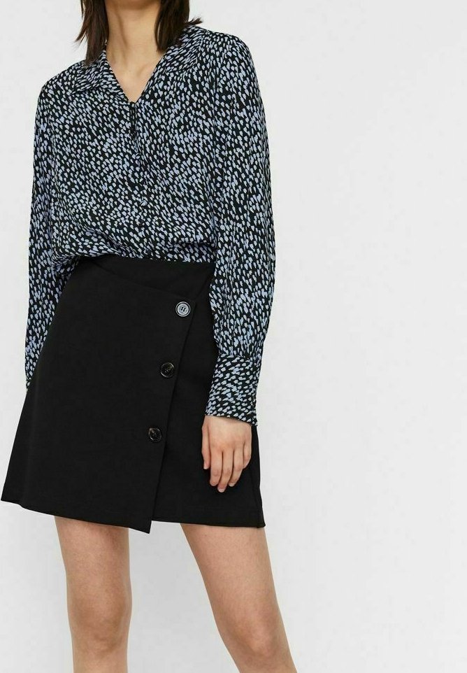 Femme portant une mini-jupe noire avec trois boutons et un chemisier à manches longues noir et blanc à motifs, debout devant un fond uni.