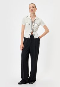 Maglietta bianca a righe con maniche corte, rifinita con bordi e bottoni neri, abbinata a pantaloni neri larghi e scarpe nere; design semplice e moderno.