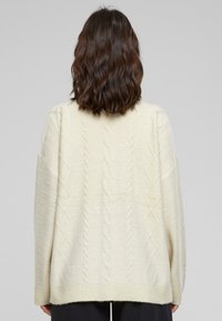 Cremfarbener gestrickter Pullover mit einem Zopfmuster, weiten Ärmeln und einem gerippten Saum. Die Rückansicht hebt das strukturierte Design hervor.