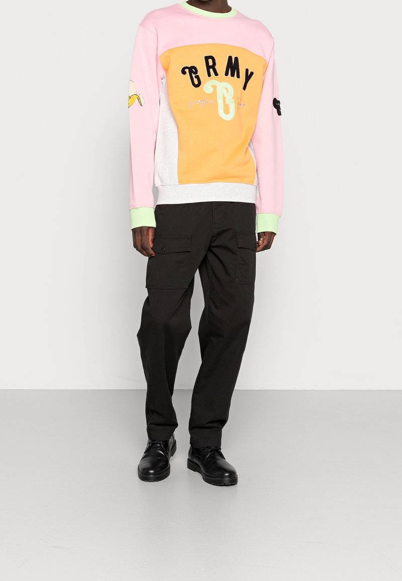 Personne portant un sweat-shirt rose et orange à manches longues avec le texte "GRMY", un pantalon cargo noir et des chaussures noires, debout devant un fond uni.