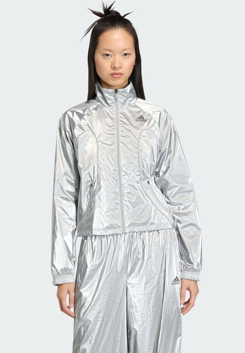 Veste de sport argentée avec col montant, fermeture éclair et panneaux en maille respirante. Comprend des poignets élastiques et un pantalon assorti argenté.