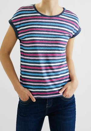 Osoba ubrana w koszulkę z krótkim rękawem w wielokolorowe poziome paski oraz ciemnoniebieskie jeansy, z rękami w przednich kieszeniach.