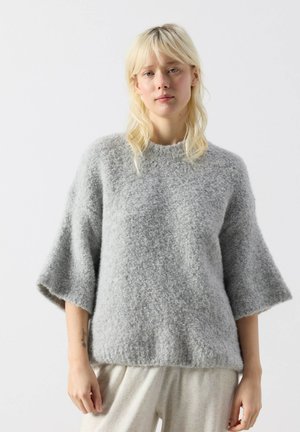 Młoda kobieta o blond włosach, ubrana w luźny, szary, teksturowany sweter z szerokimi rękawami do łokcia i jasne spodnie, stojąca na białym tle.