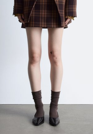 Jambes portant une jupe à carreaux marron, des chaussettes marron retroussées et des chaussures noires à bouts pointus, debout sur un sol gris face à un mur blanc.