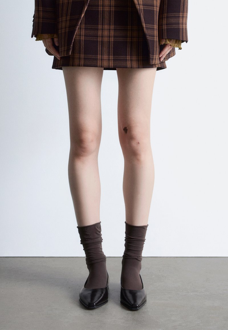 Jambes portant une jupe à carreaux marron, des chaussettes marron retroussées et des chaussures noires à bouts pointus, debout sur un sol gris face à un mur blanc.