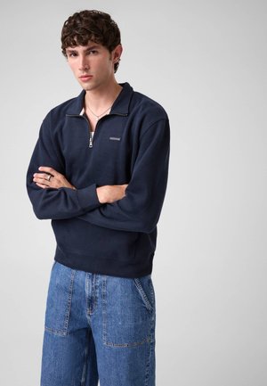 Junger Mann mit kurzem lockigem Haar, der einen marineblauen Sweatshirt mit Viertelreißverschluss und blaue Jeans trägt, mit verschränkten Armen vor einem schlichten Hintergrund.