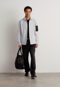 Cămașă gri cu guler, tricou negru și blugi închiși la culoare. Cămașa are un buzunar cu un accent texturat. Poartă o geantă închisă la culoare.