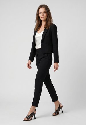 Kvinde går selvsikkert iført sort blazer, sorte skræddersyede bukser, hvid bluse og sorte sandaler med hæl mod en lys, ensfarvet baggrund.