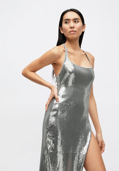 Strieborné flitrové šaty s priliehavým strihom, tenkými ramienkami a vysokým rozparkom. Hladká textúra s reflexným povrchom a jednoduchým výstrihom.