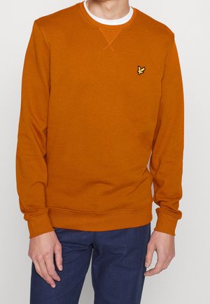 Mand iført en rustorange crewneck-sweatshirt med lille gul og sort fuglelogo på brystet, kombineret med blå ternede bukser.