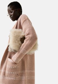 Black woman wearing large glasses and a textured brown and white coat, holding a beige fluffy handbag under her arm.
