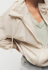 Veste beige clair à fermeture éclair avec capuche, en tissu texturé et léger. Portée sur un t-shirt vert clair à col en V et un jean clair.