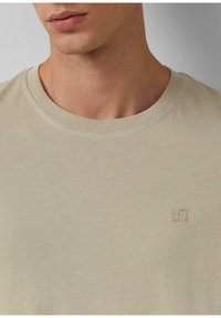 Zbliżenie na osobę w beżowej koszuli z okrągłym dekoltem z małym, wypukłym logotypem "s.Ol" na lewej piersi.