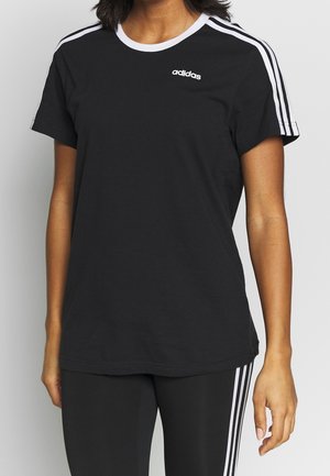 Kobieta nosząca czarną koszulkę Adidas z białymi paskami na ramionach oraz dopasowane czarne spodnie z białymi paskami.