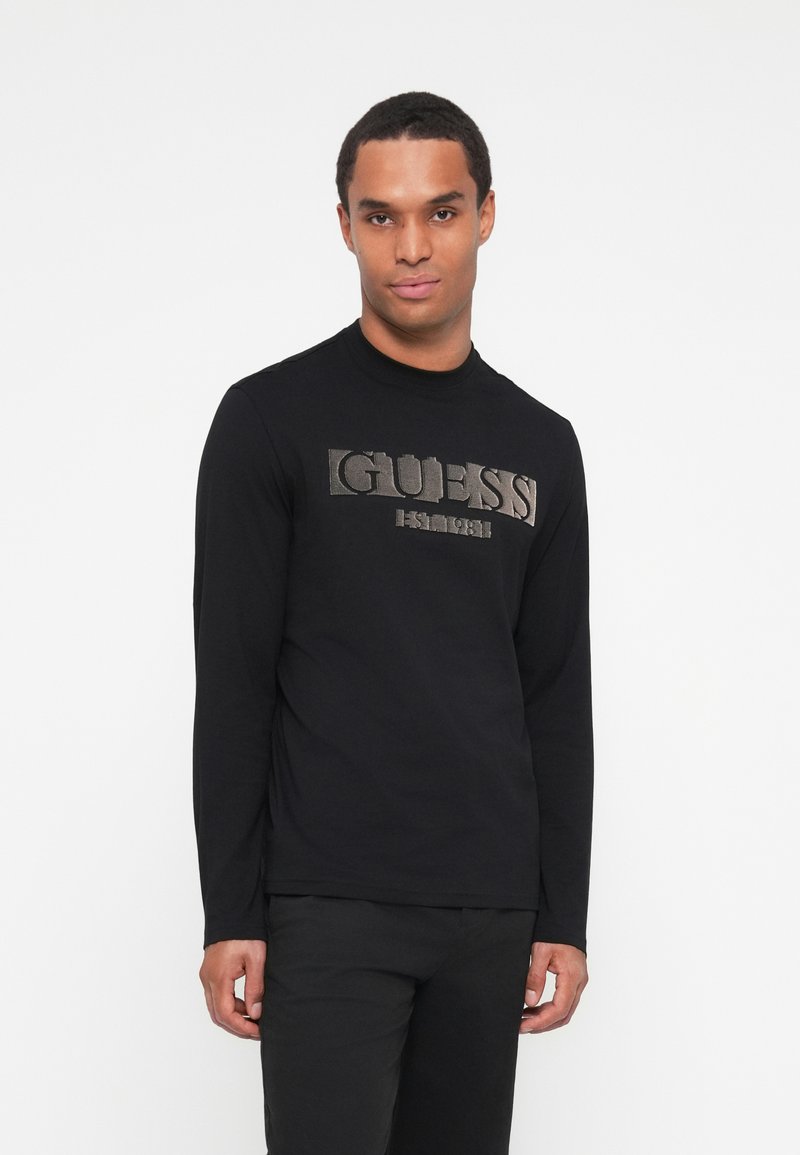 Svart langermet skjorte med et metallisk sølv "GUESS"-logo foran, med rund hals og en glatt, tettsittende tekstur.