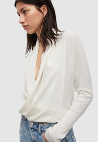 Weißes, geripptes Langarmoberteil mit einem drapierten V-Ausschnitt, das eine weiche Textur und enganliegende Bündchen hat, kombiniert mit hellblauen Jeans.