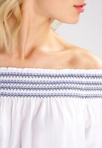 Haut blanc à épaules dénudées avec encolure smockée et broderie en zigzag bleu et noir ; tissu doux, léger avec une finition texturée.