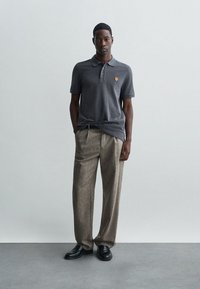 Γκρι πόλο μπλούζα με ένα μικρό κεντημένο λογότυπο, συνδυασμένη με ανοιχτό καφέ, πιεσμένες παντελόνες και μαύρα παπούτσια. Απλή, καθημερινή εμφάνιση.