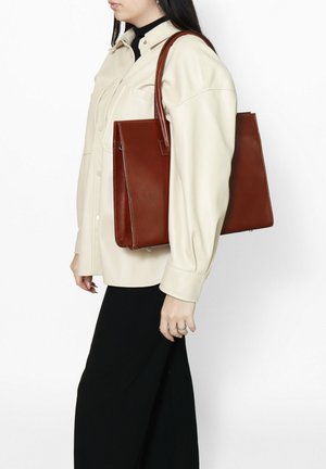 Braune Leder-Tote-Bag, an der Seite gehalten, mit einem strukturierten Design, glatter Textur und verlängerten Tragegriffen. Kombiniert mit einem hellbeigen Oberteil.