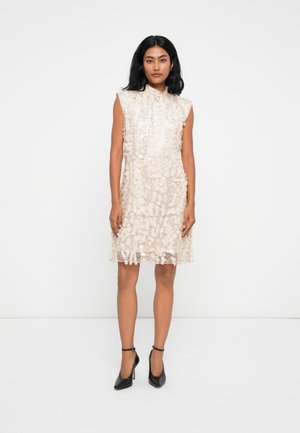 Robe en dentelle beige clair avec un col montant et une coupe sans manches, ornée de motifs floraux et d'un ourlet festonné. Assortie à des escarpins noirs.