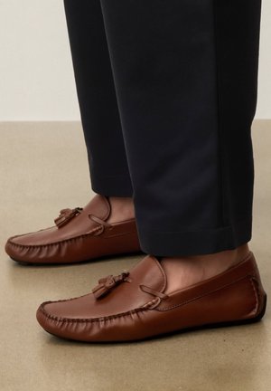 Brune læder tasselloafers båret med mørke, afkortede bukser på en beige gulv.