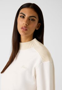 Kremowy sweter z teksturowanym, prążkowanym, beżowym kołnierzem. Posiada dyskretną grafikę z przodu oraz luźny krój. Gładki materiał.