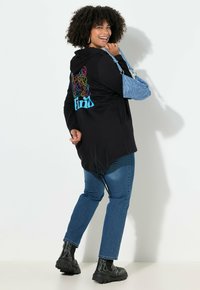 Mujer sonriente con cabello rizado que lleva una chaqueta con capucha negra con un estampado colorido en la parte posterior, jeans azules, botas negras y sostiene un bolso azul sobre el hombro.