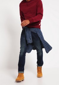 Punahkonen pyöreäkauluksinen collegepaita rintataskulla, sininen ruudullinen paita vyötärölle solmittuna, tummansiniset farkut ja keltaiset työkengät ruskeilla nauhoilla.