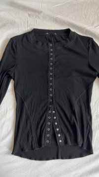 Black ribbed long-sleeve top with round neckline, front snap closure, and side seam detailing. Soft texture, slim fit design.