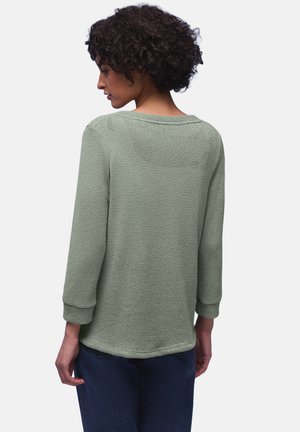 Kobieta z krótkimi, kręconymi włosami, ubrana w jasnozielony, teksturowany top z długim rękawem i ciemnoniebieskie spodnie, widziana od tyłu na jednolitym tle.