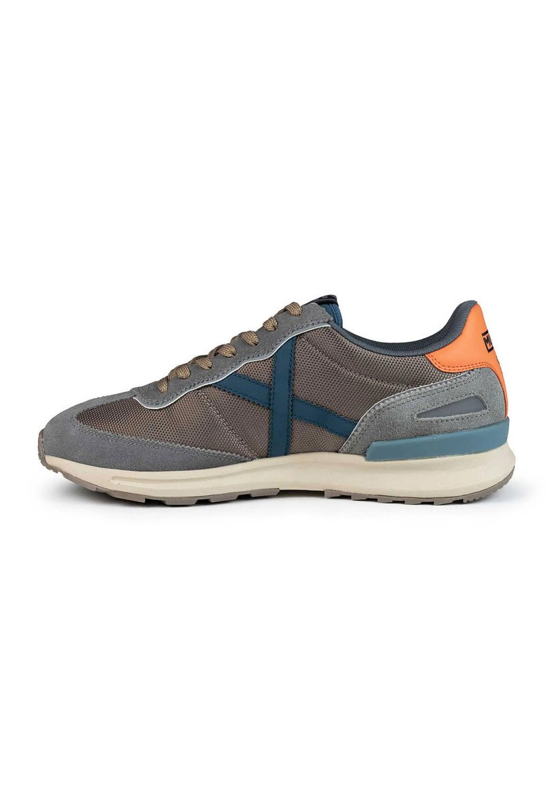 Zapatilla deportiva gris con materiales de malla y gamuza. Presenta acentos en azul, cordones beige y una lengüeta trasera naranja. Suela lisa para tracción.
