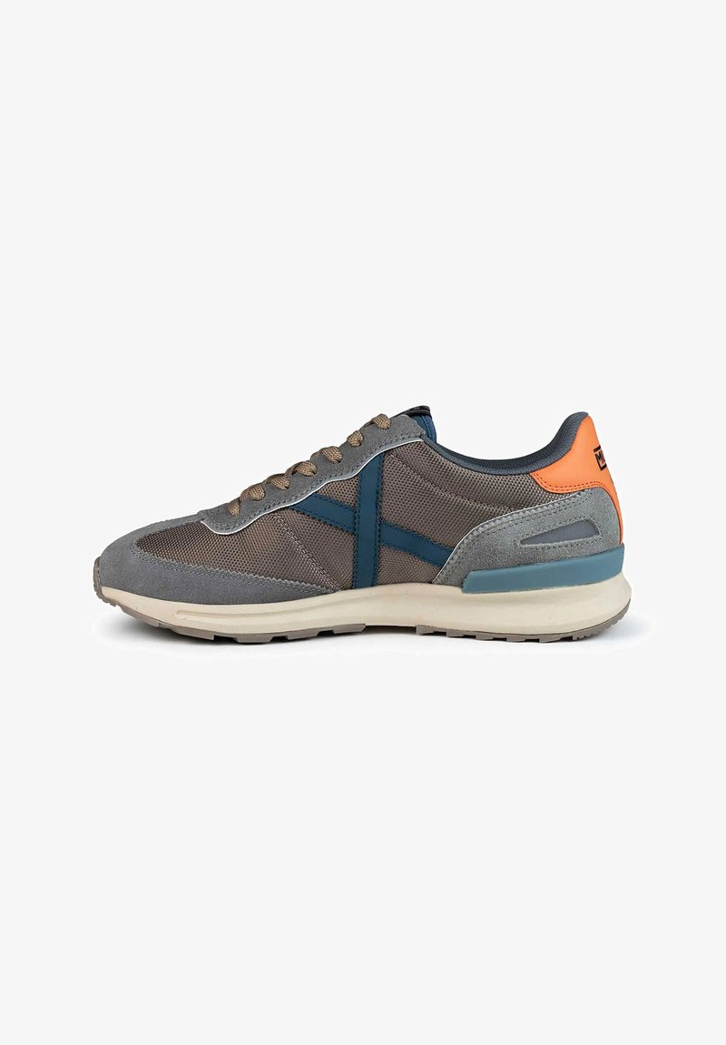 Zapatilla deportiva gris con materiales de malla y gamuza. Presenta acentos en azul, cordones beige y una lengüeta trasera naranja. Suela lisa para tracción.