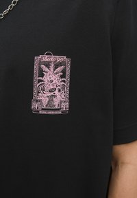 Sort t-shirt med et pink grafiktryk af et tropisk design, der inkluderer planter og teksten "studio 90", i et blødt bomuldsstof.