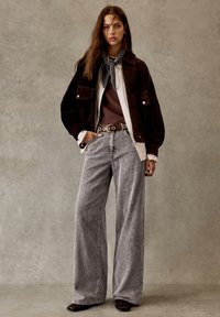 Bruine suede jas, witte shirt, donker sweater, grijze corduroy wijde broek, zilveren hardware op de riem, sjaal om de hals.