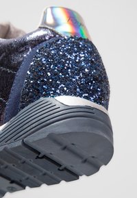 Primo piano di un tacco di sneaker in tessuto glitter blu scuro e una suola in gomma nera testurizzata con un dettaglio iridescente lucido sulla parte superiore.