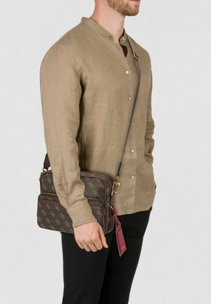 Hombre con camisa beige abotonada y pantalones negros, llevando un bolso bandolera marrón con un patrón de monograma y una etiqueta de cuero roja.