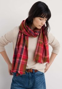 Ternet tørklæde i rød, brun og grøn, lavet af stof med frynsete kanter. Modellen bærer det draperet over en lysefarvet trøje.