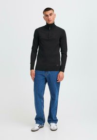 Svart zip-neck tröja, stickad tyg, åtsittande design, ribbad nederkant och ärmslut. Kombinerad med blå jeans och grå/vita sneakers.